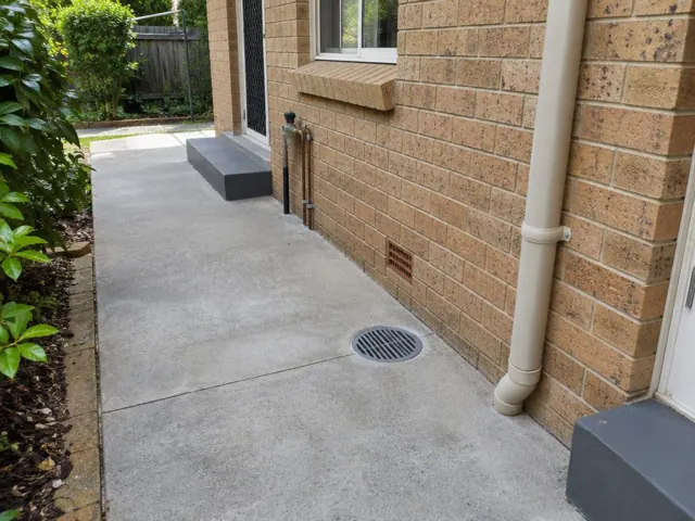 Outdoor gully drain with grate set in a concrete path beside a brick house in Turramurra