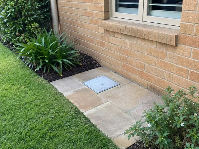 Square access cover flush in tiled paving beside a brick wall and lawn in Wahroonga